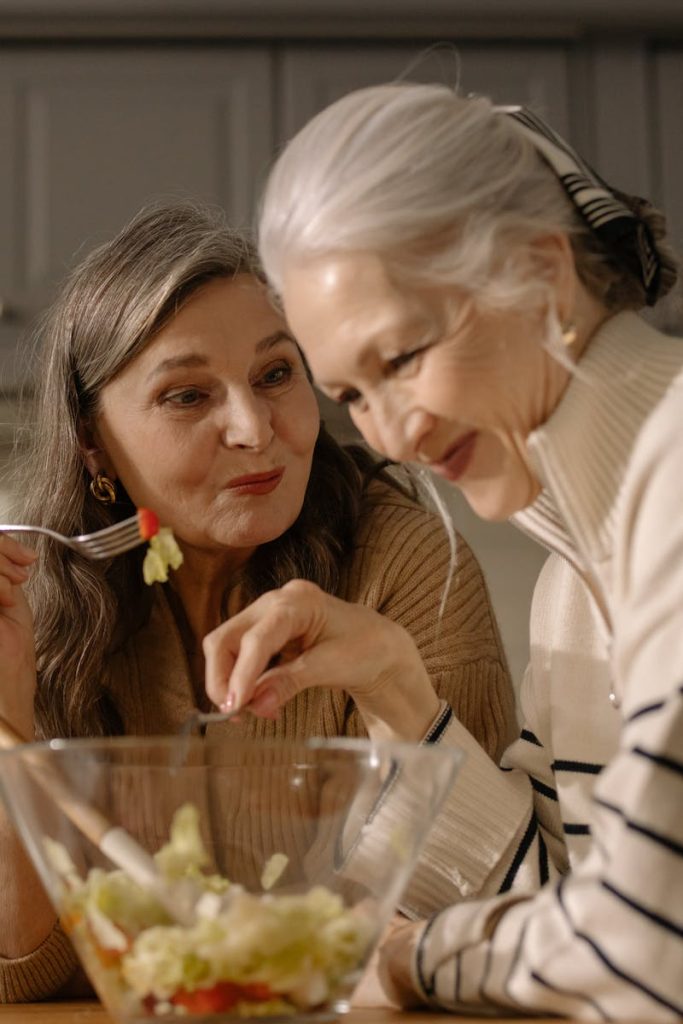 Two senior women smiling and enjoying a fresh salad in a cozy kitchen setting.